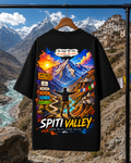 A black short-sleeve t-shirt on a wooden hanger, displaying a colorful graphic print of Spiti Valley. The background is a real outdoor mountain landscape with snow-capped peaks and a river.